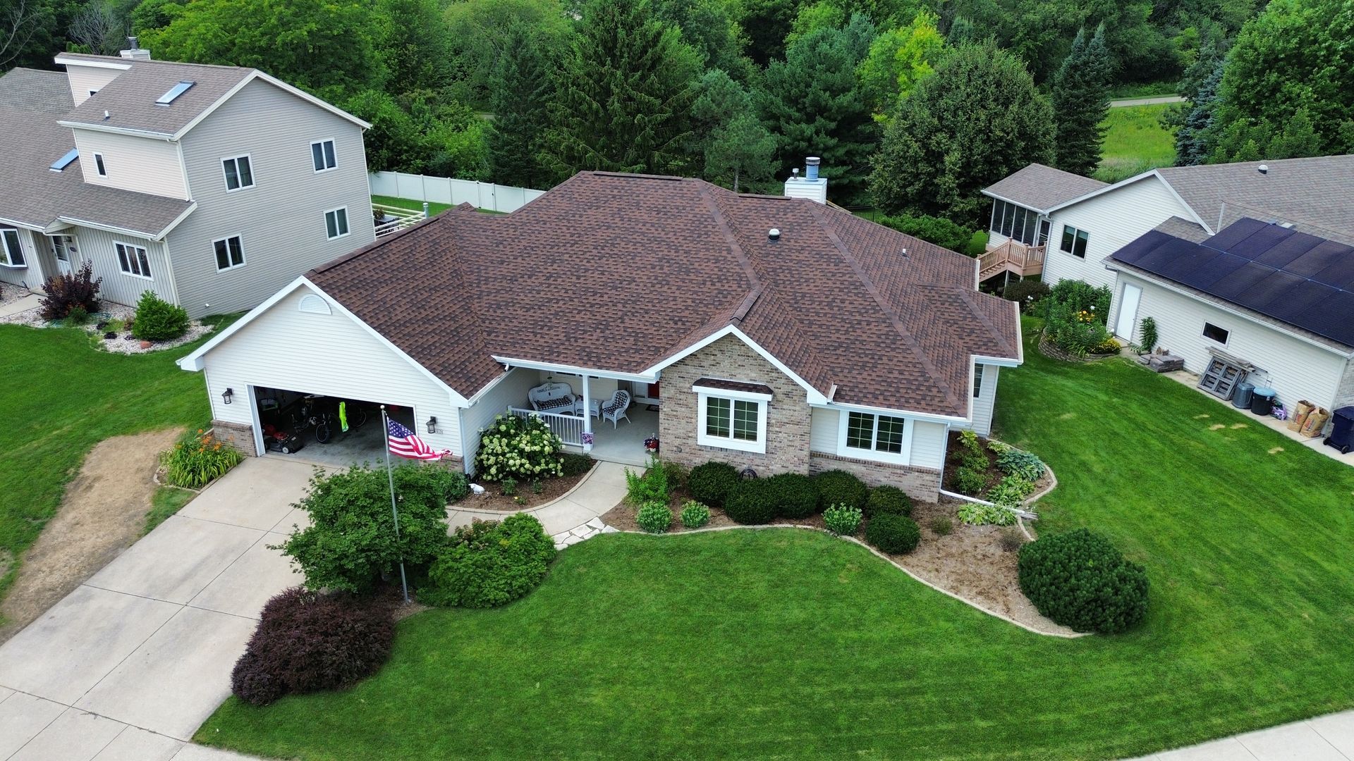 An aerial view of a suburban house with brown roofing, white siding, and a landscaped front yard on a sunny day.