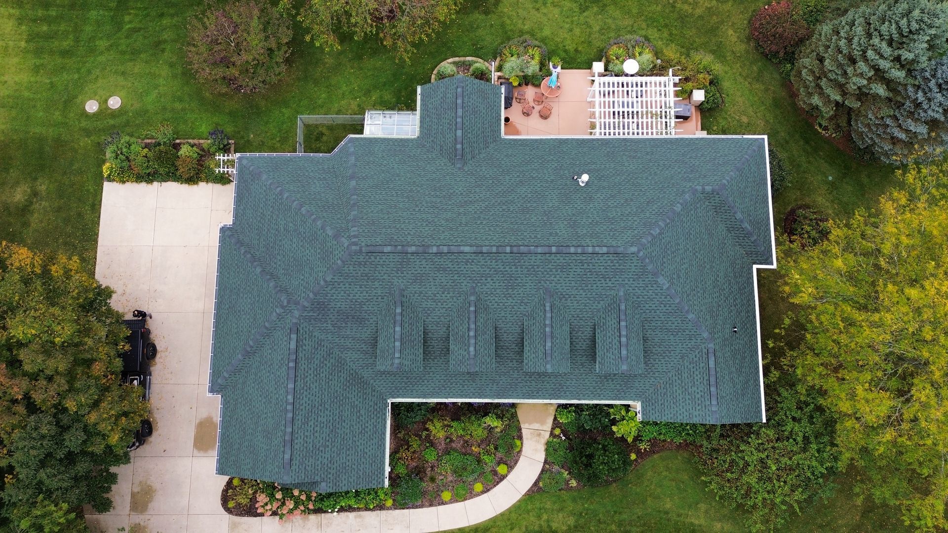 A top-down aerial view of a dark-shingled house with four front-facing dormers, surrounded by trees and a paved driveway.