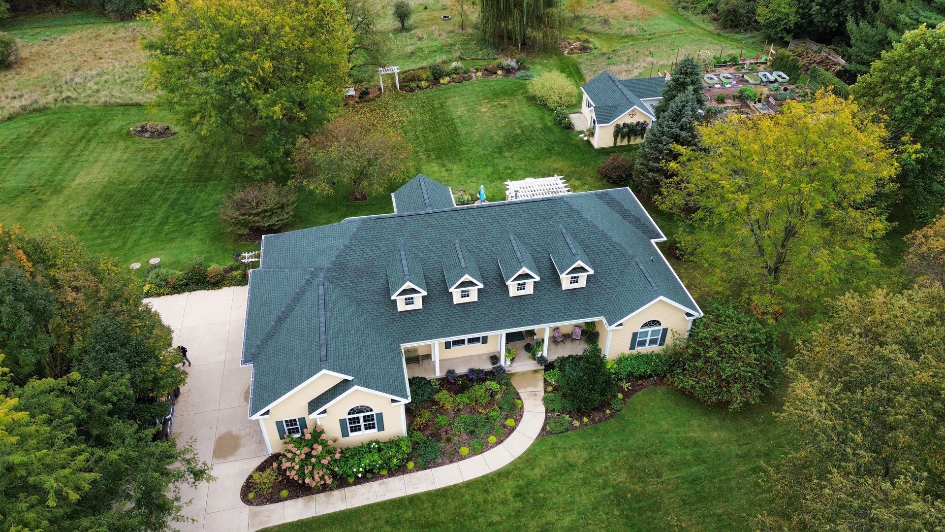 An aerial view of a tan, single-story suburban house with a dark green roof, surrounded by lush trees and a green lawn.
