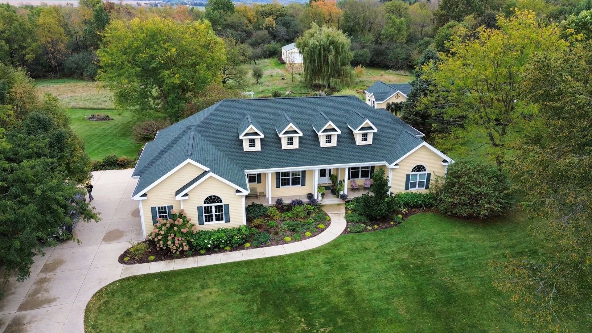 A light-colored house with a dark roof and four dormer windows, set in a green, tree-lined landscape with a curved walkway.
