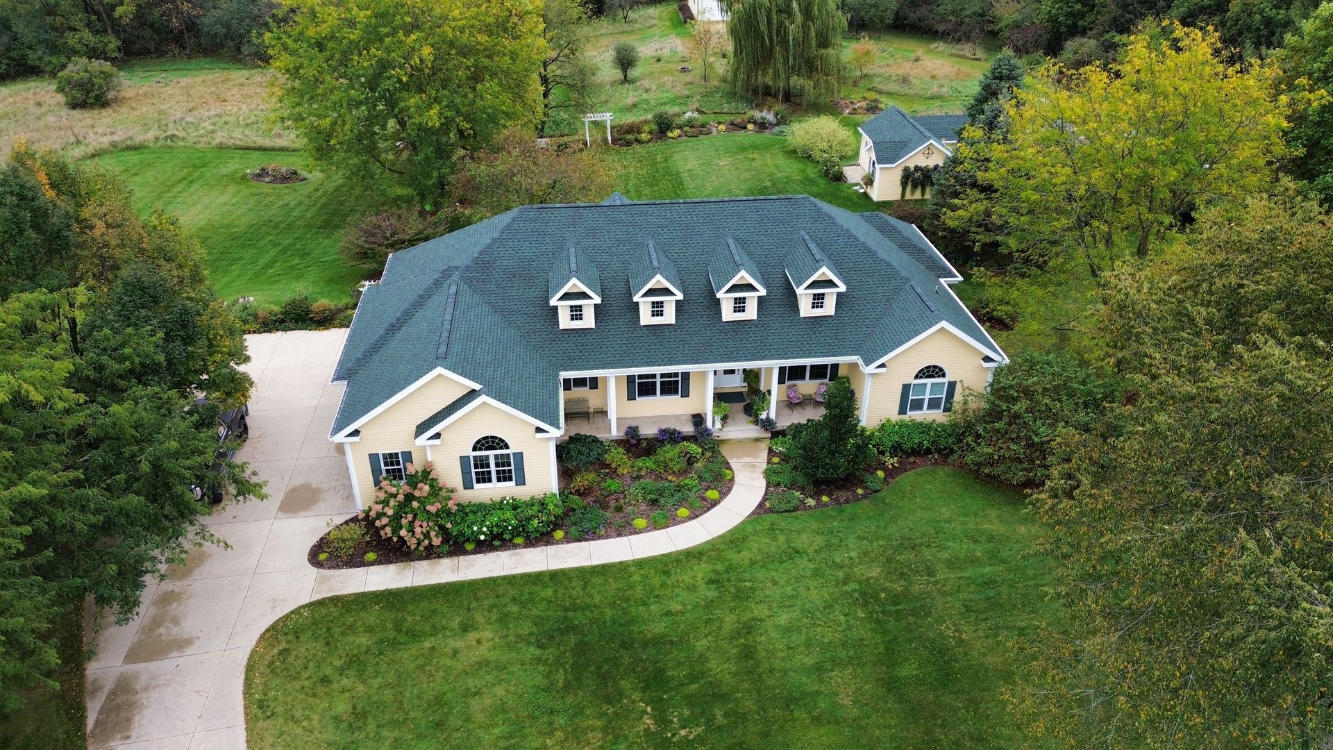 An aerial view of a tan, ranch-style house with a dark green roof, surrounded by a green lawn and trees.