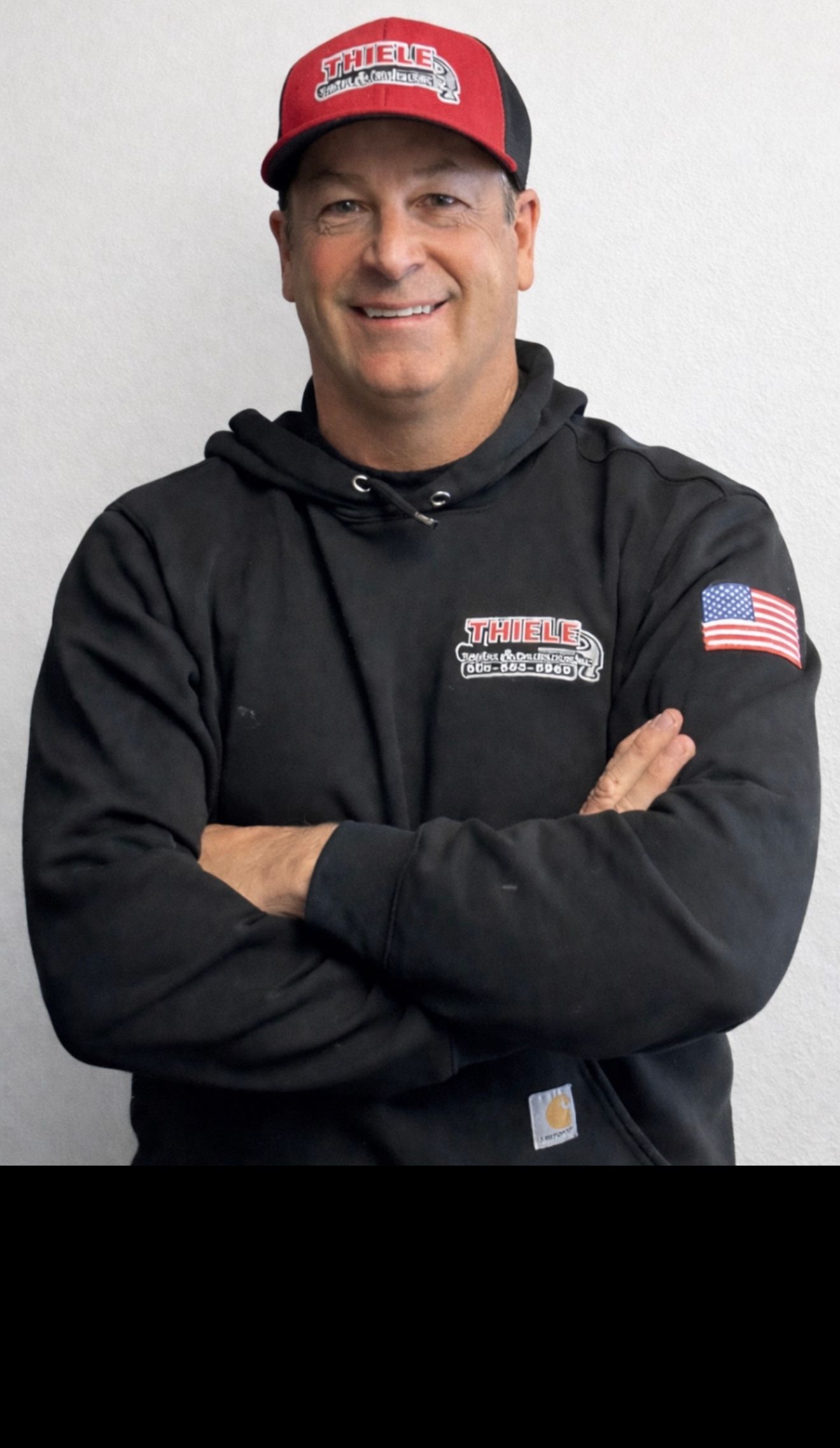 A person wearing a black hoodie with an American flag patch and a red cap, arms crossed, smiling against a white background.