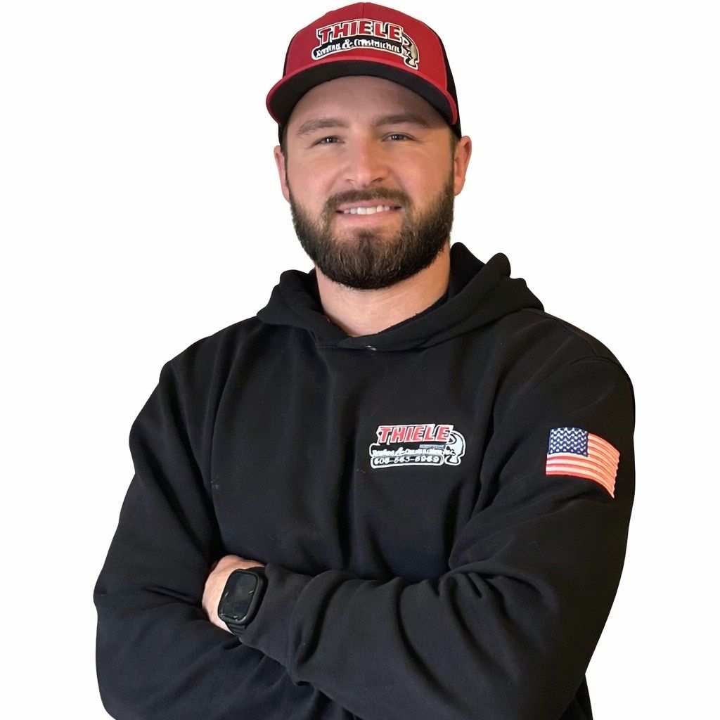 A smiling person wearing a red and black baseball cap and a black hoodie with an American flag patch, arms crossed.