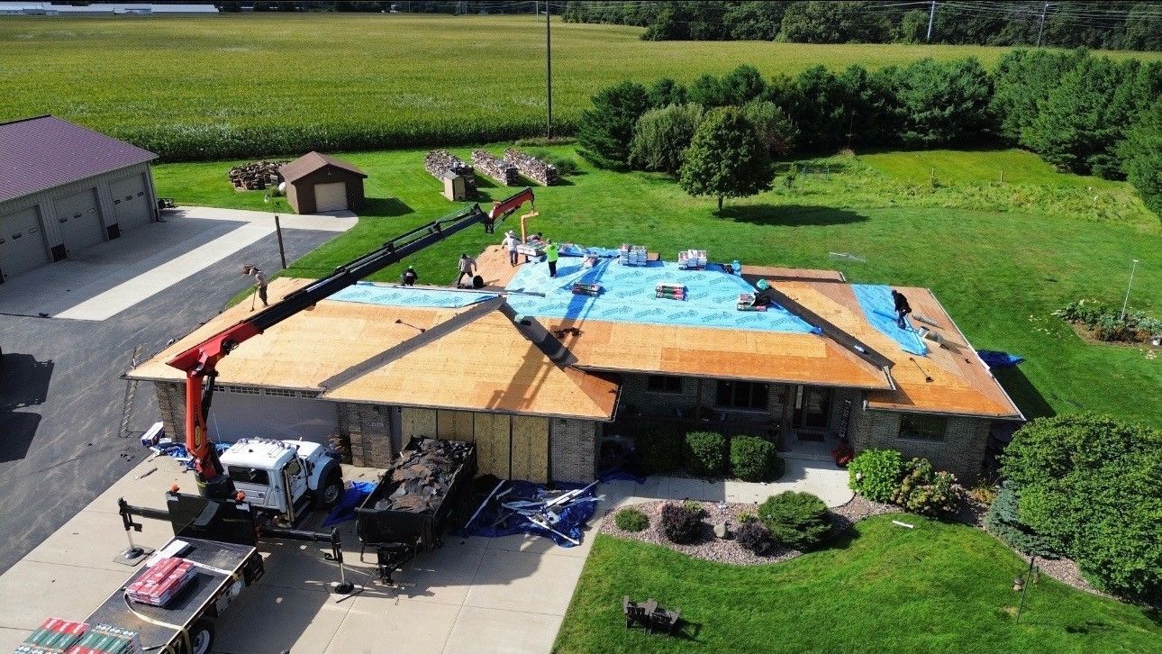 A crane lifts supplies to a house with a partially exposed wooden roof and a blue tarp, set in a rural landscape.