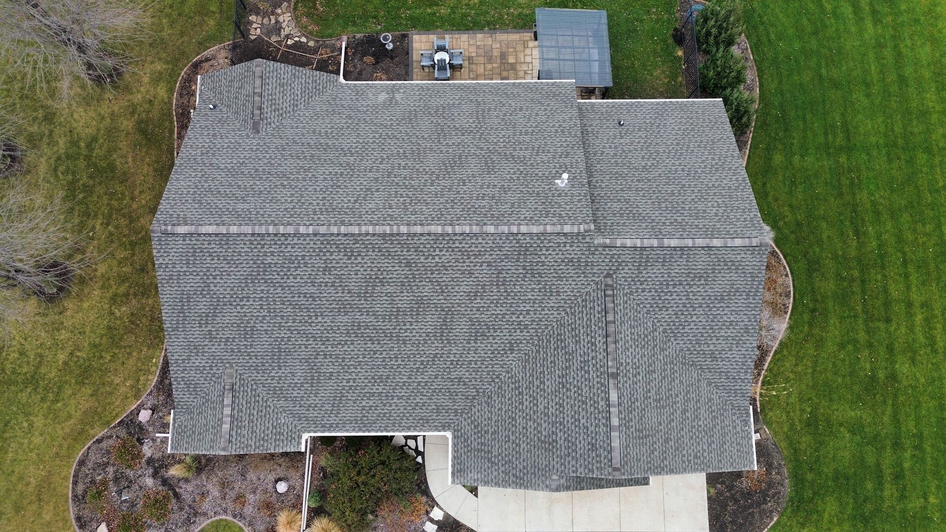 A top-down aerial view of a gray shingled roof on a house, surrounded by lawn and landscaping.
