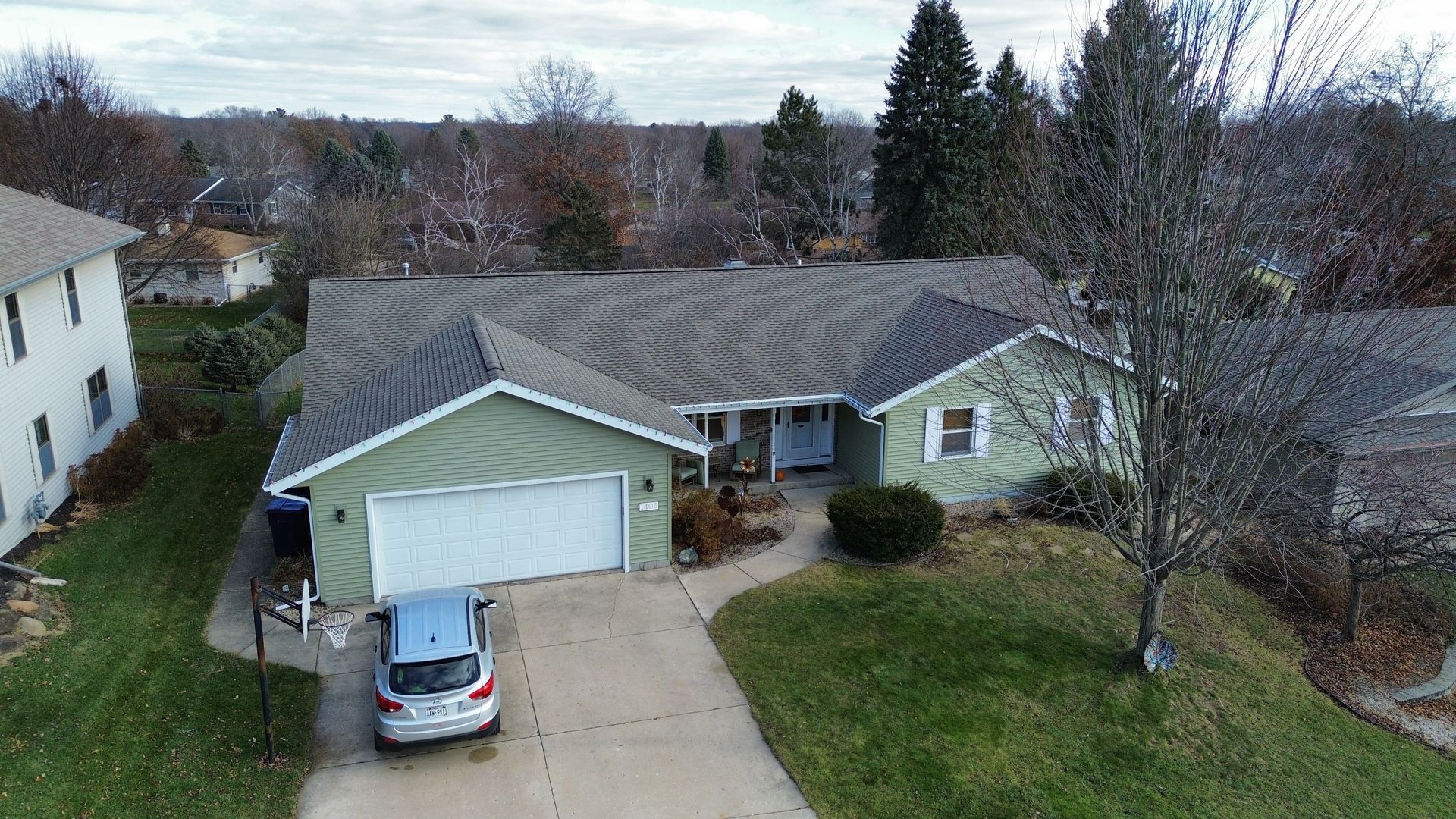 An aerial view of a light green suburban house with a two-car garage, a light-colored driveway, and bare trees in autumn.