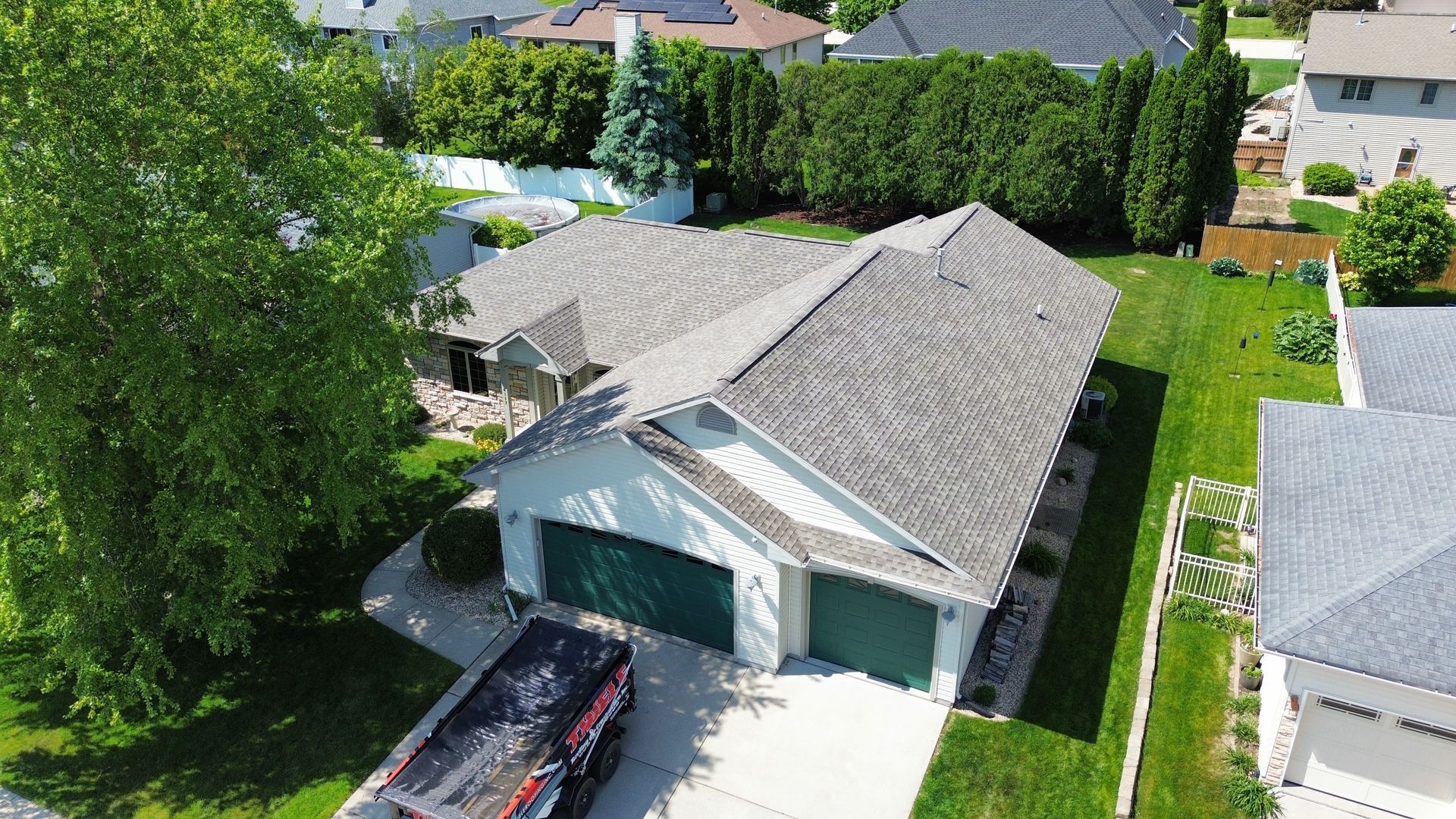 Aerial view of a residential house with a grey roof, dark green garage doors, and a dumpster in the driveway.
