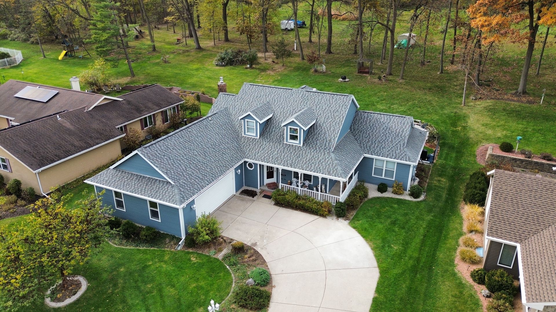 An aerial view of a blue, single-story house with a grey shingled roof, a curved driveway, and surrounding green lawn.