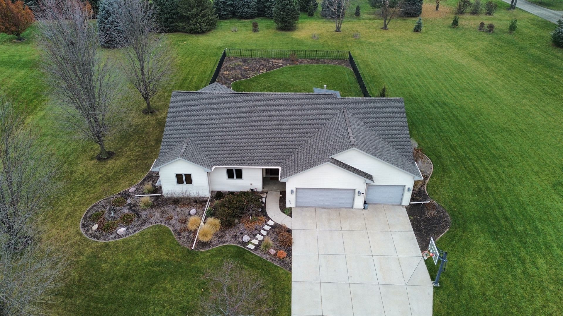 An aerial view of a single-story house with a light-colored exterior, a grey roof, and a concrete driveway in a green yard.