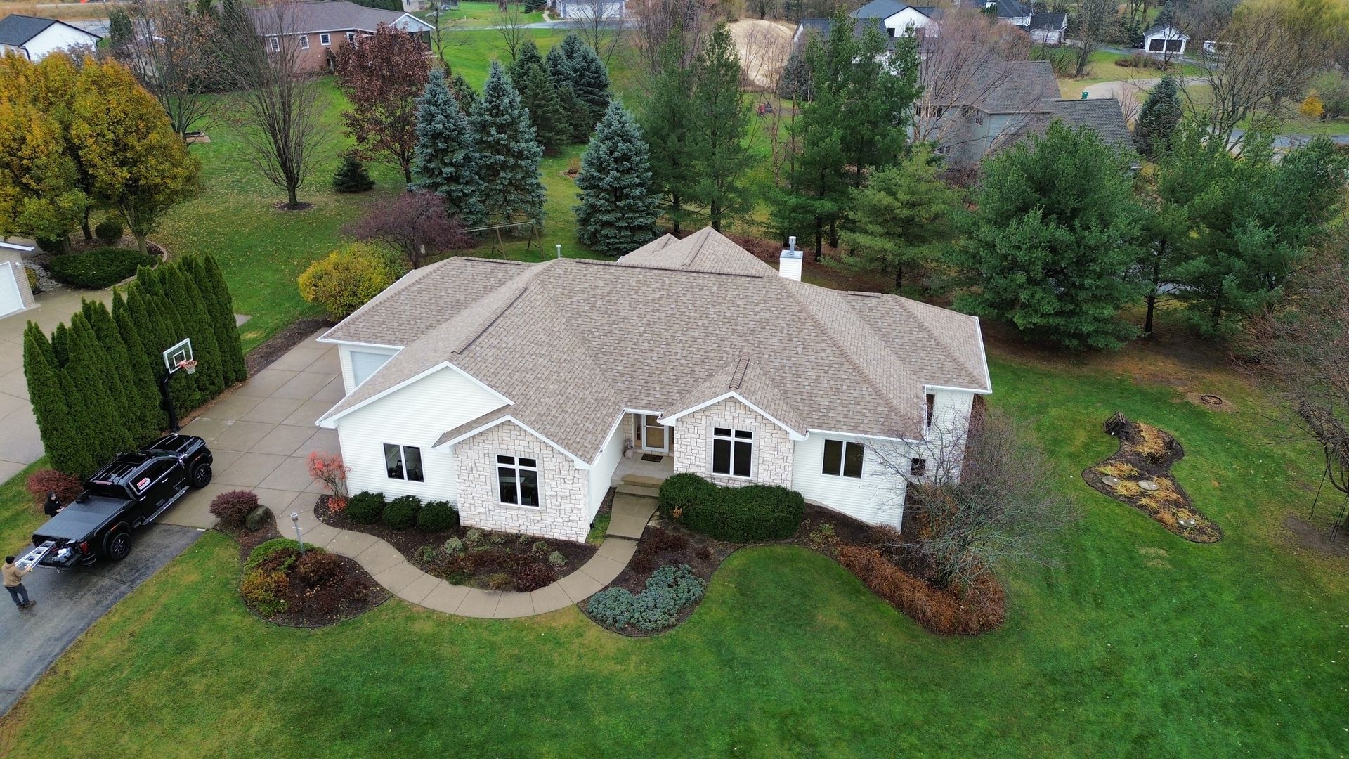 An aerial view of a light-colored, single-story suburban house with a brown roof, surrounded by a lawn and evergreen trees.