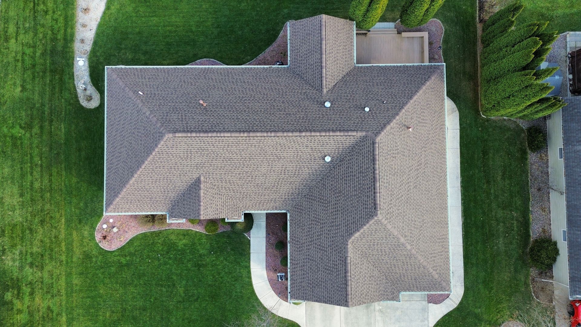 An aerial view of a single-story residential house with a gray shingle roof, surrounded by green grass and landscaping.
