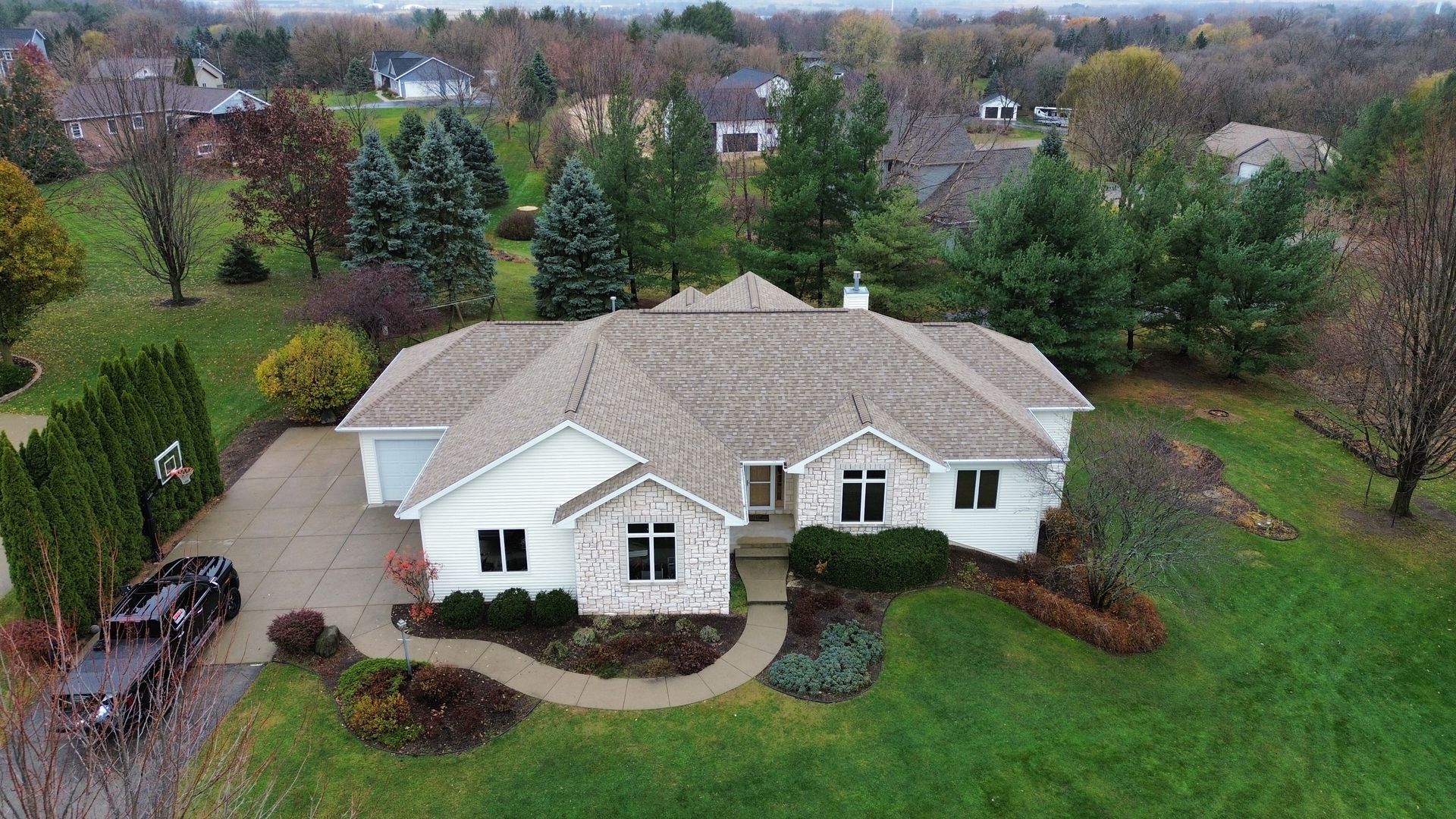 An aerial view of a single-story white house with a tan shingled roof, a paved driveway, and lush green lawns.