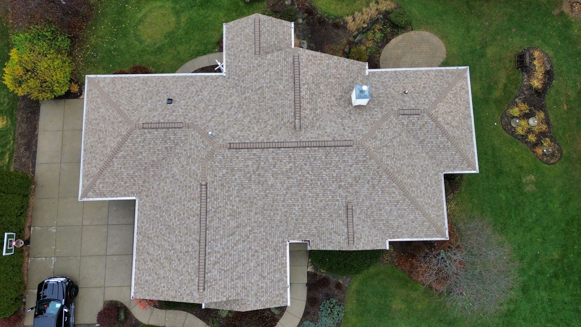 An aerial view of a tan, multi-pitched residential roof surrounded by a green lawn, walkways, and a parked car.