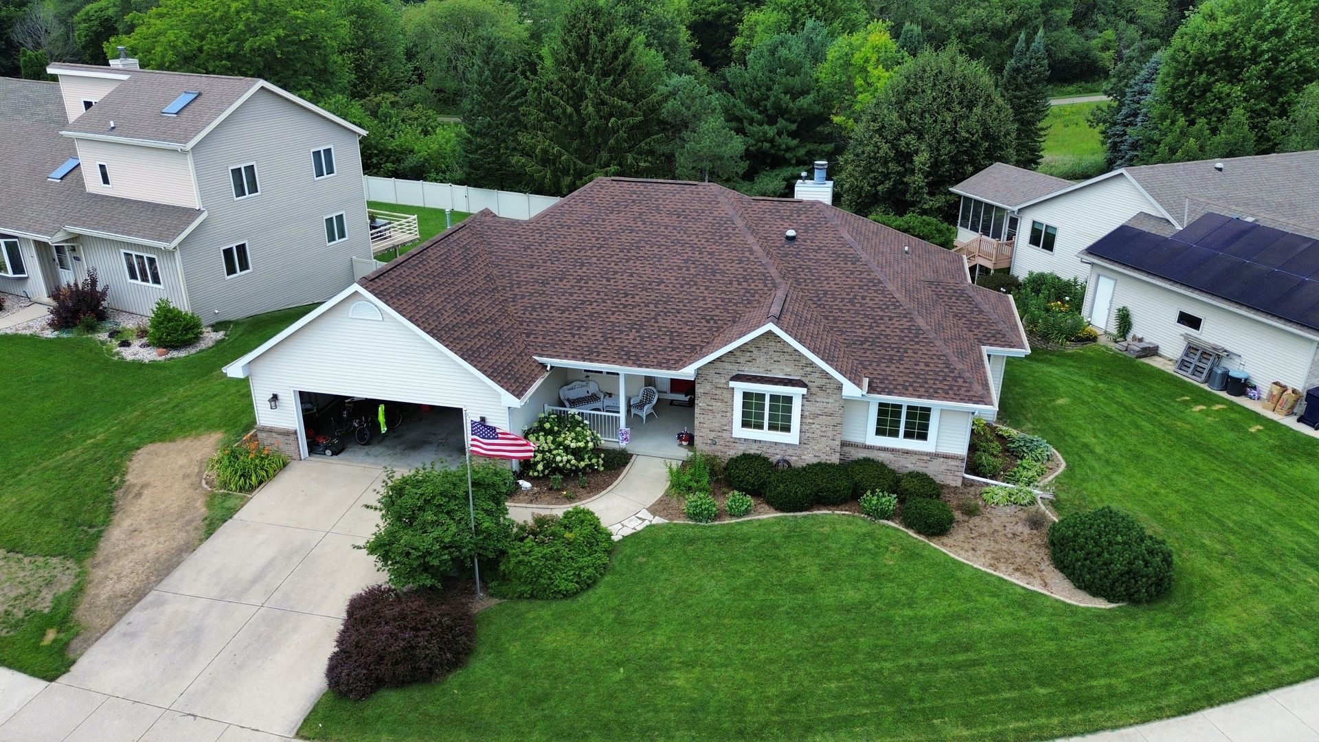 Drone view of a brown-roofed house with white trim, a driveway, and a well-manicured lawn surrounded by trees.