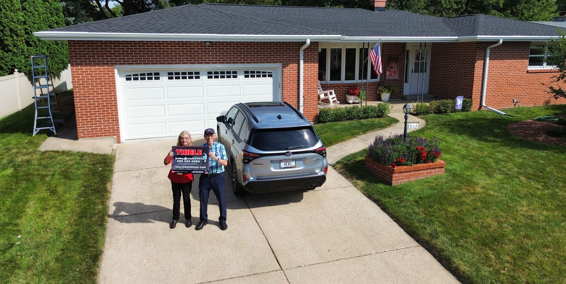 Two people stand in a brick home's driveway holding a sign next to a silver SUV, with a ladder leaned against the house.