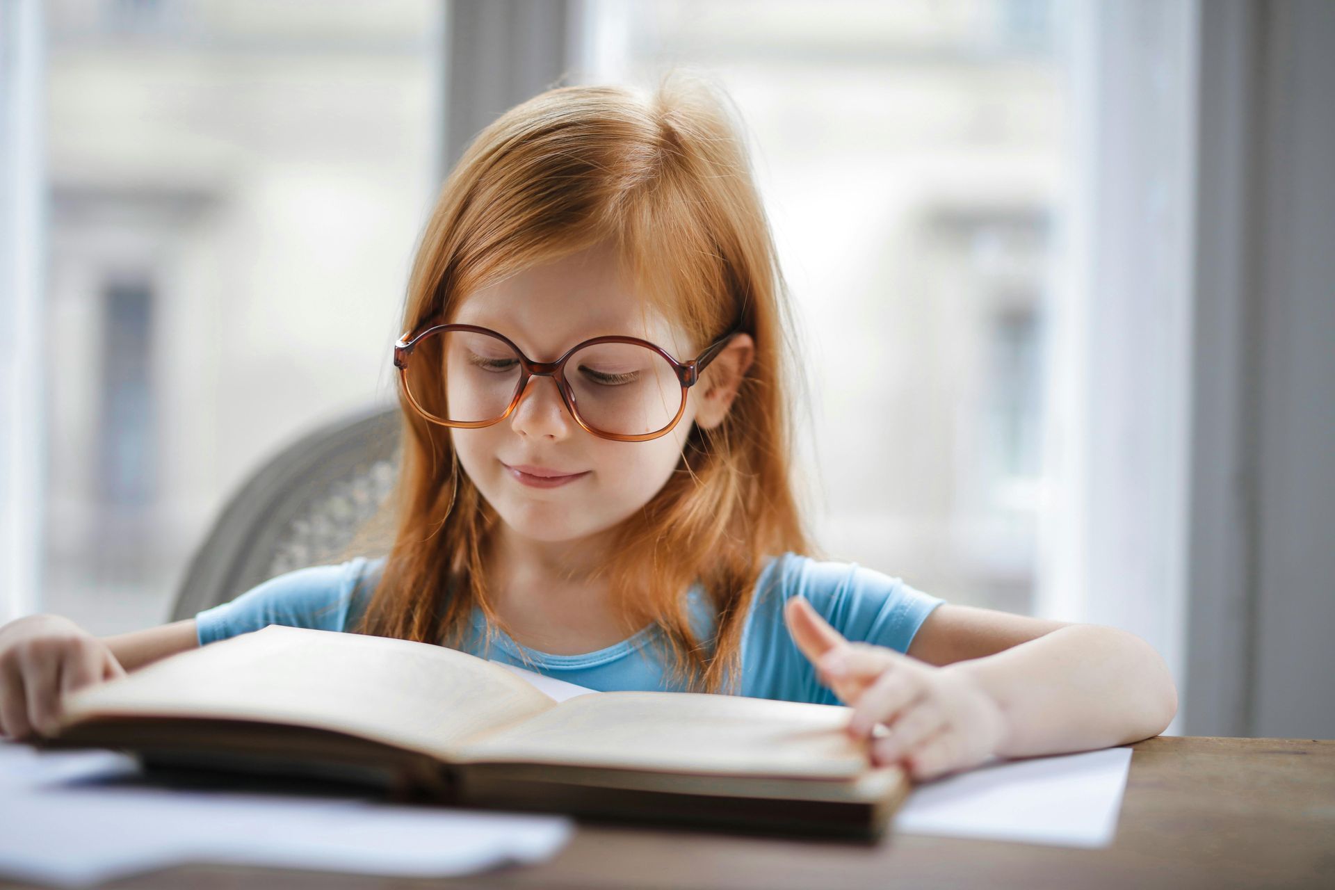 A little girl wearing glasses is reading a book at a table.
