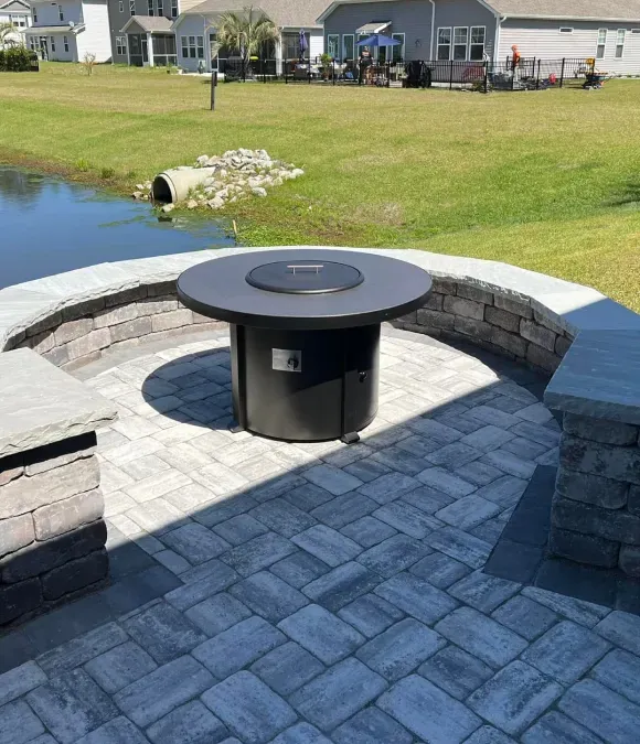 Backyard patio with a circular fire pit beside a pond, surrounded by stone seating and paver walkway