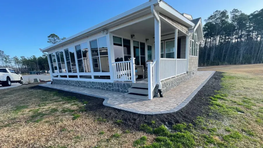 White house with wraparound porch and ramp on a grassy lot, with trees in the background