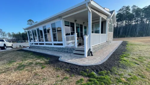 White house with wraparound porch and ramp on a grassy lot, with trees in the background