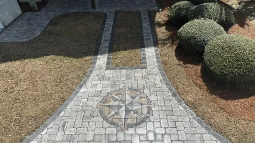 Stone pathway with a circular compass mosaic, bordered by grass and trimmed shrubs in a garden.