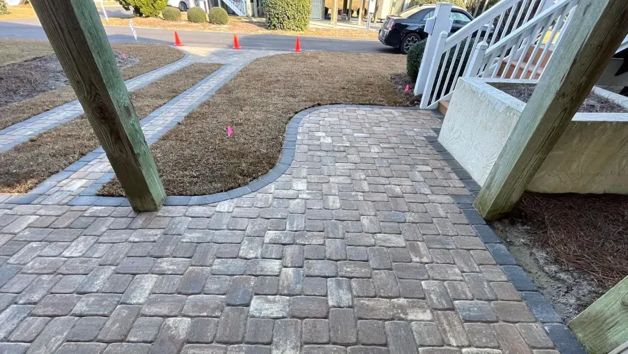 Brick walkway between wooden posts leading to a white porch beside a yard and driveway