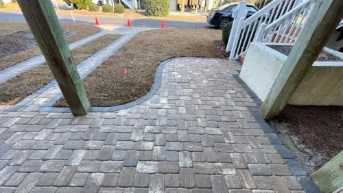 Brick walkway between wooden posts leading to a white porch beside a yard and driveway