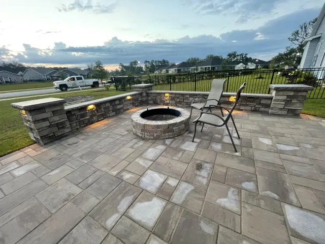 Stone patio with a lit fire pit and two chairs overlooking a lake at dusk.