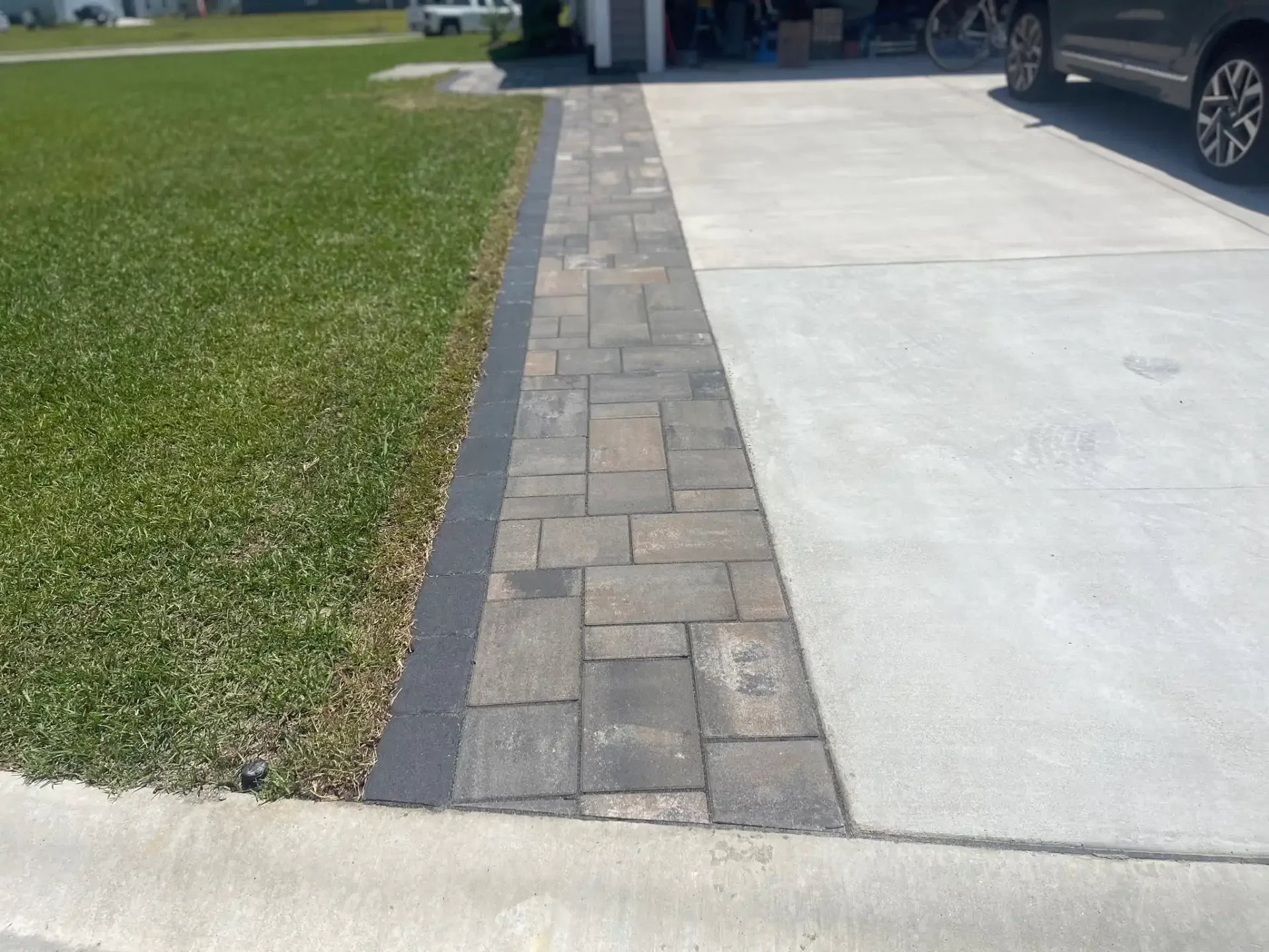 Brick paver walkway between green lawn and concrete driveway beside a parked car
