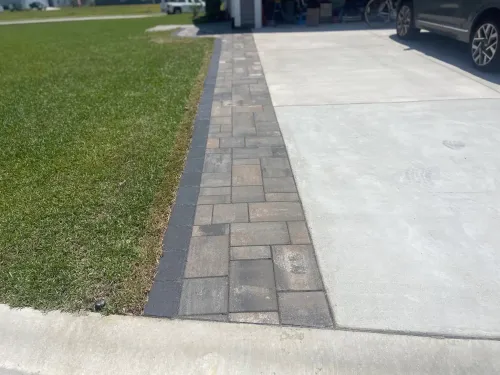 Driveway with a narrow brick paver strip between green lawn and light concrete, leading to a carport.