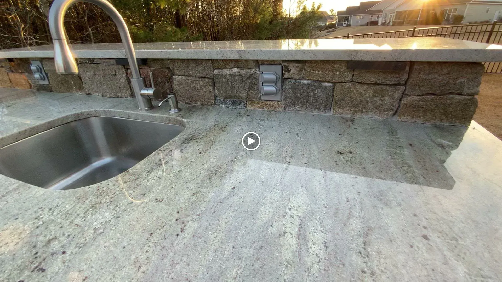 Outdoor stone countertop with a stainless steel sink, faucet, and power outlet by a low wall.