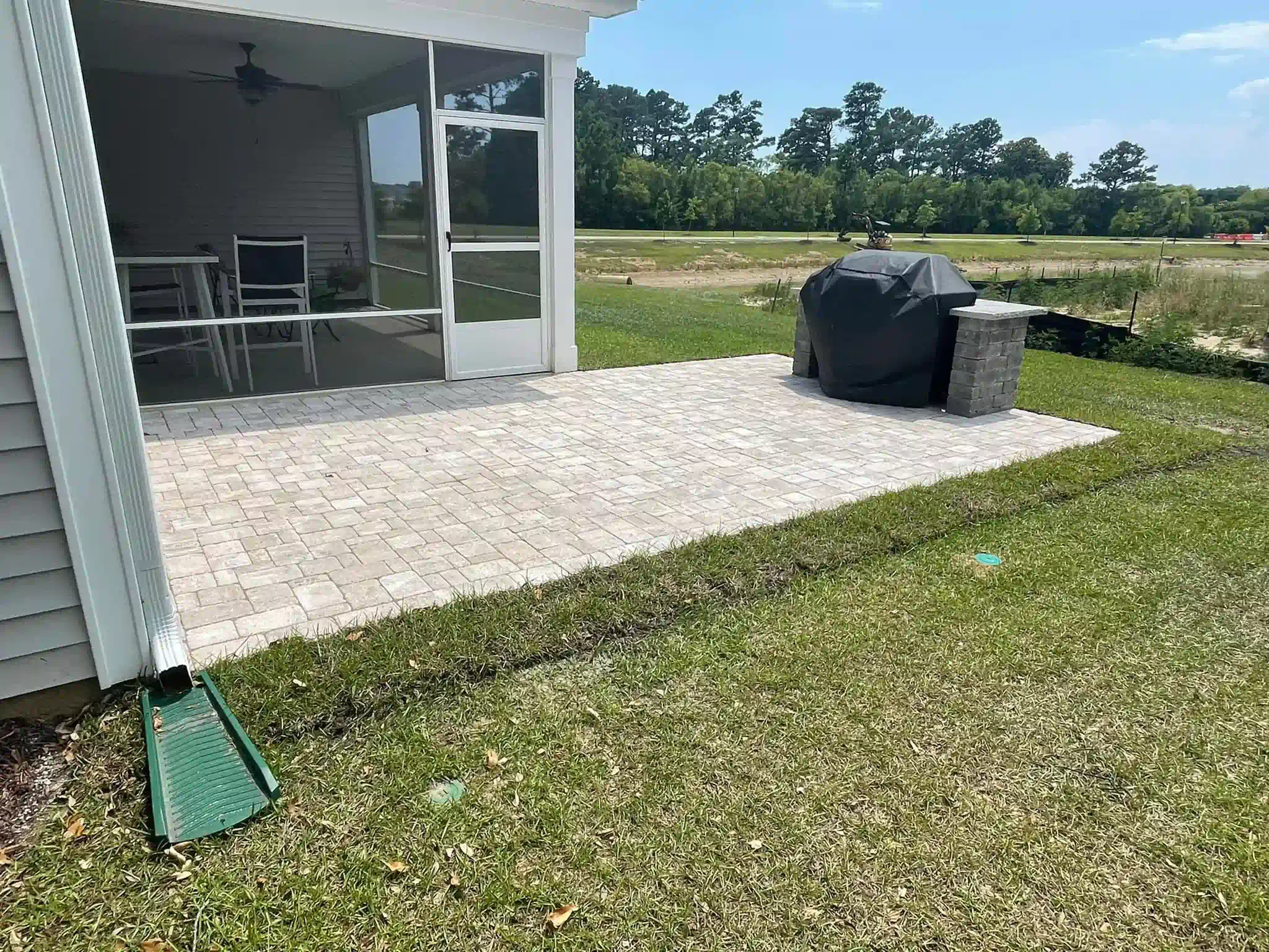 Patio with outdoor dining set and grill beside a grassy yard on a sunny day