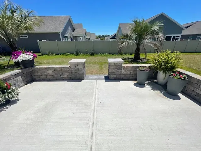 Patio with stone planters, potted flowers, and a small palm facing a grassy backyard and houses beyond.
