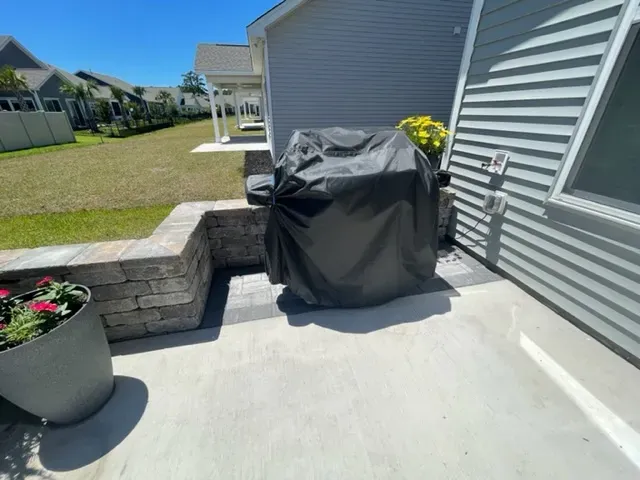 Covered grill on a concrete patio beside a gray house and stone planter wall