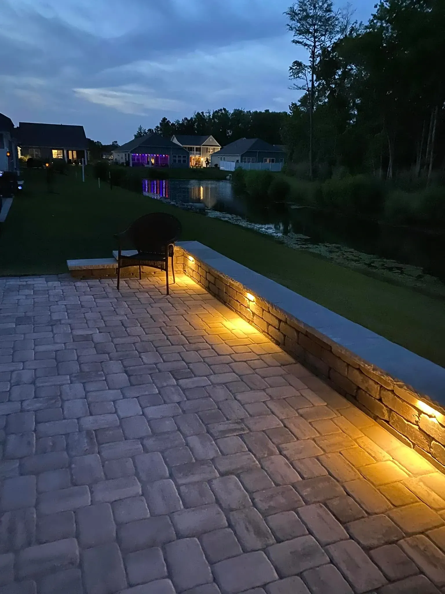 Dusk patio with warm path lights along a stone wall beside a grassy yard and houses in the distance