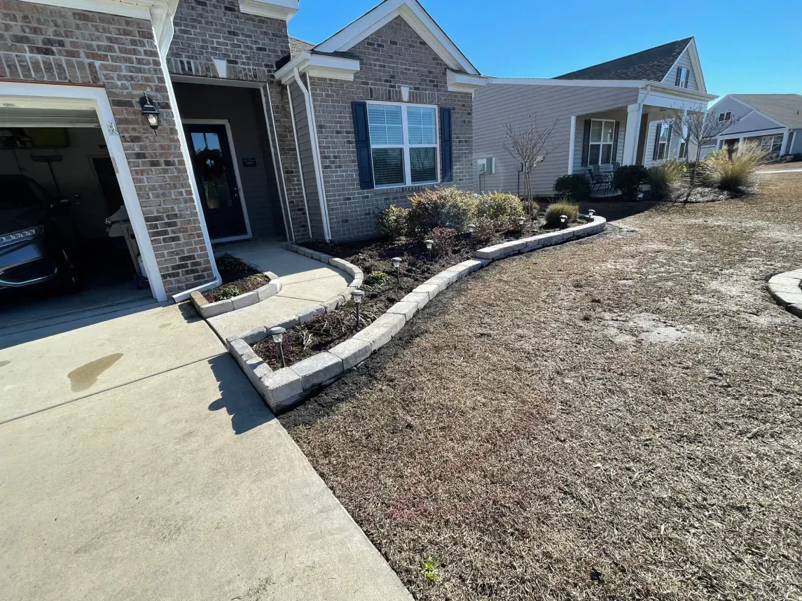 Front yard of a suburban house with a curved stone flower bed and dry lawn beside a driveway.