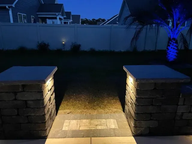 Backyard path at dusk between stone pillars, with blue-lit palm and a fence in the background