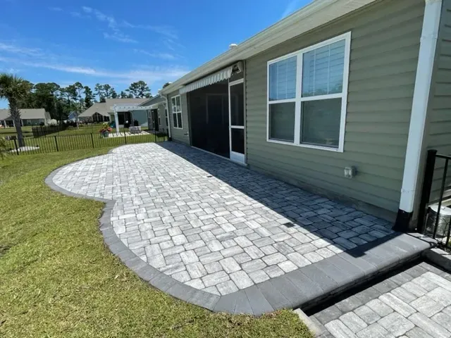 Curved gray paver patio beside a light green house with screened door and windows, bordered by grass