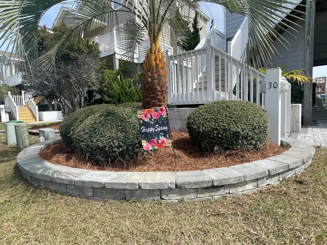 Front yard landscaping with a palm tree, shrubs, and a decorative sign by a white porch and driveway gate