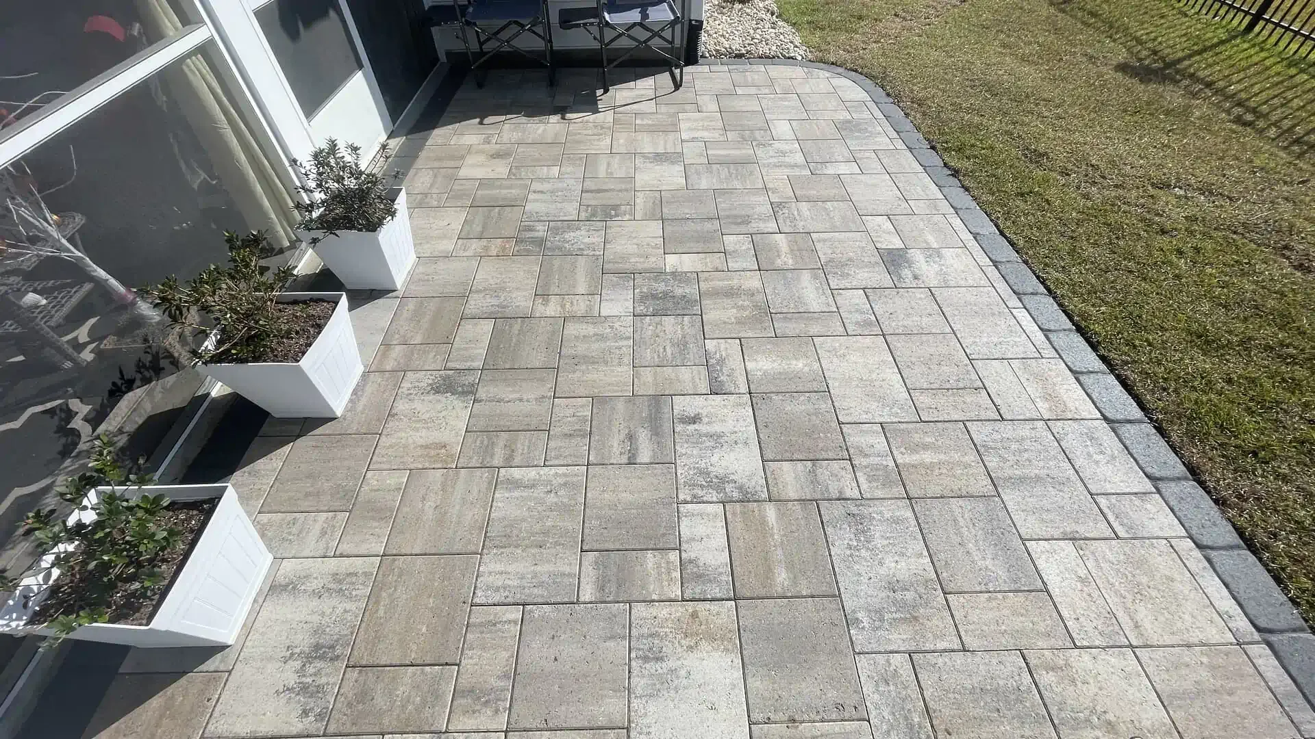 Stone paver walkway beside a house and grassy yard, leading past small planters and outdoor furniture.