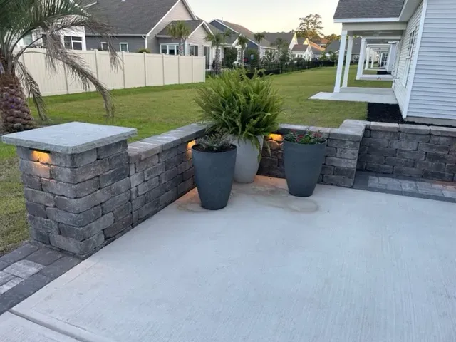 Back patio with gray pavers, stone half-wall, and two large potted plants overlooking a fenced lawn