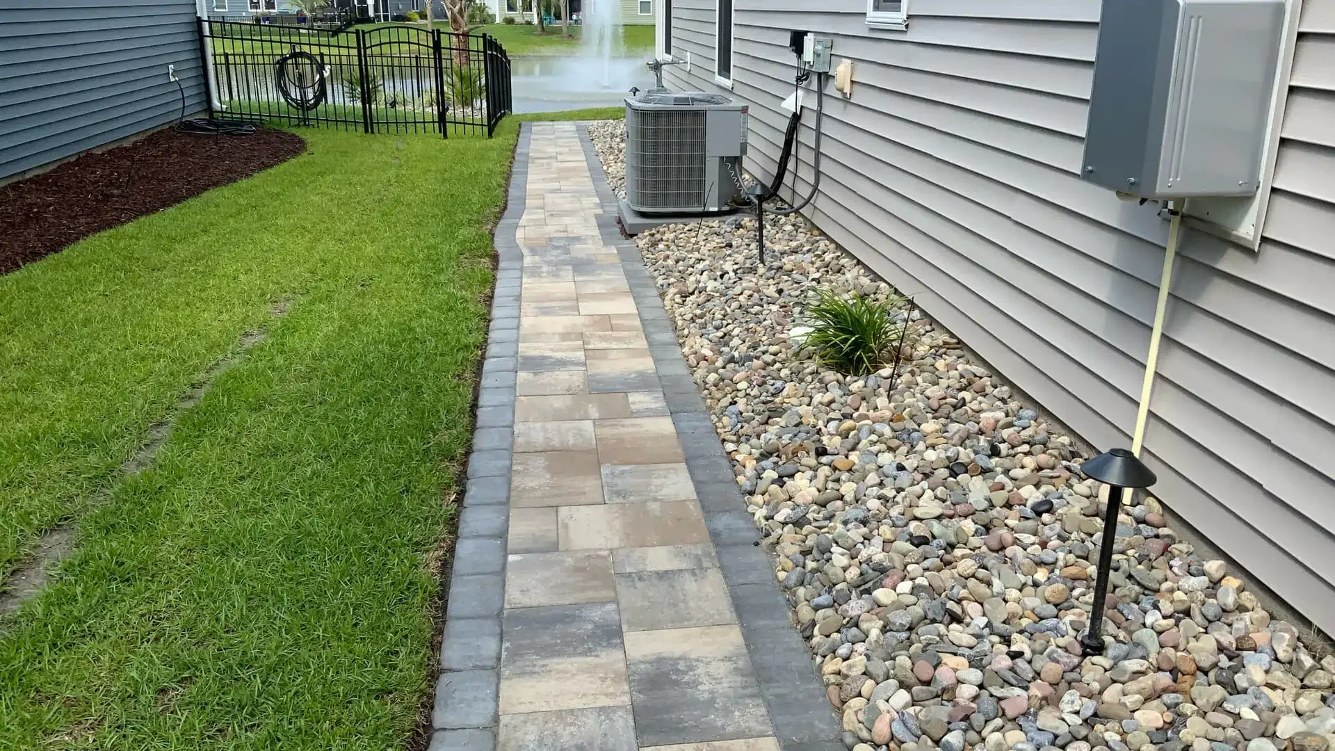 Backyard side path with paver walkway, green lawn, house siding, gravel strip, and air conditioner unit