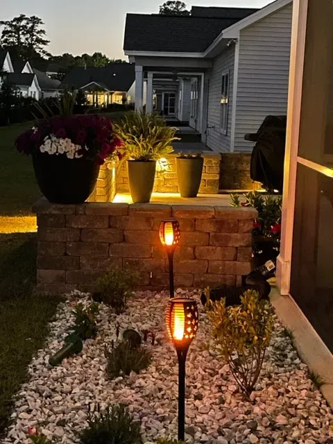 Nighttime front yard with lit tiki torches, white rocks, potted plants, and a stone retaining wall.