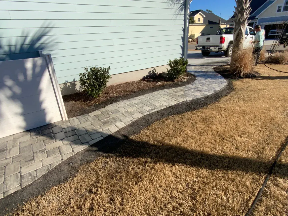Curved stone walkway beside a light blue house, with brown grass and shrubs in front yard.