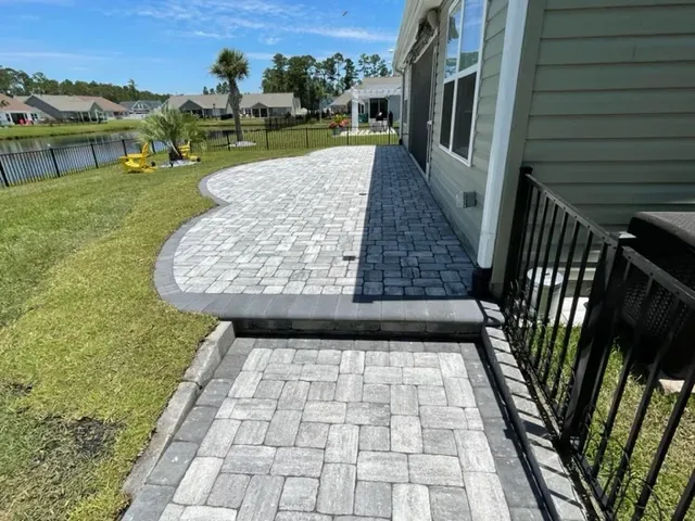 Curved gray paver walkway beside a house and canal under a sunny blue sky