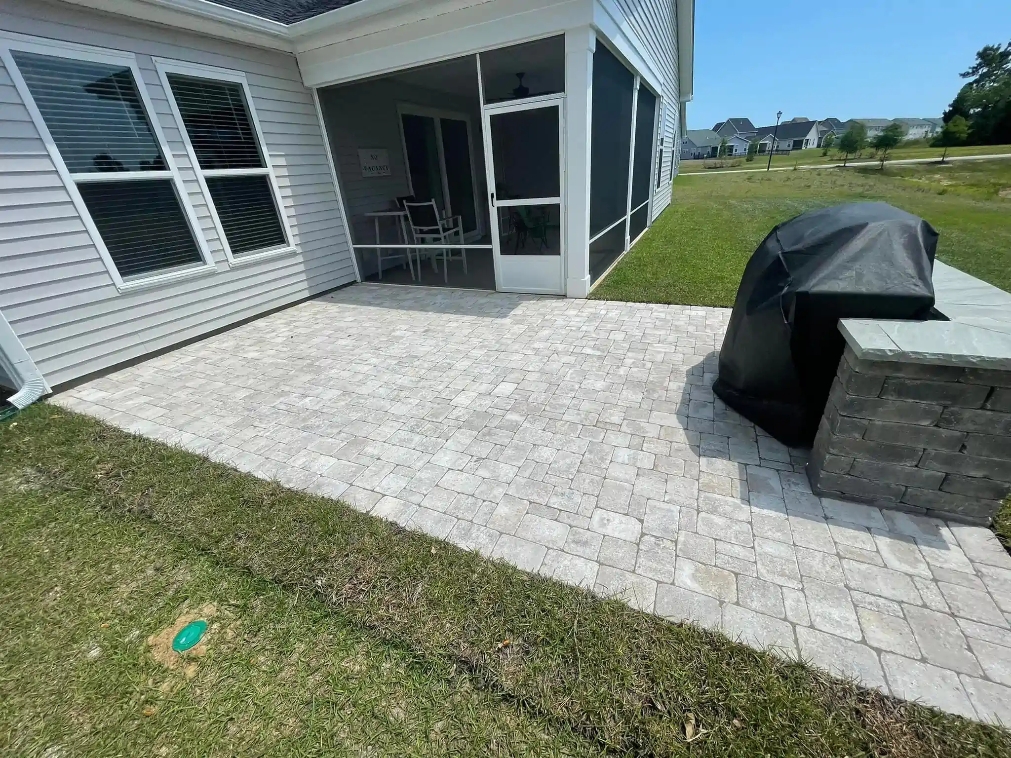 Patio with white pavers beside a gray house, covered grill, and grassy yard under a screened porch