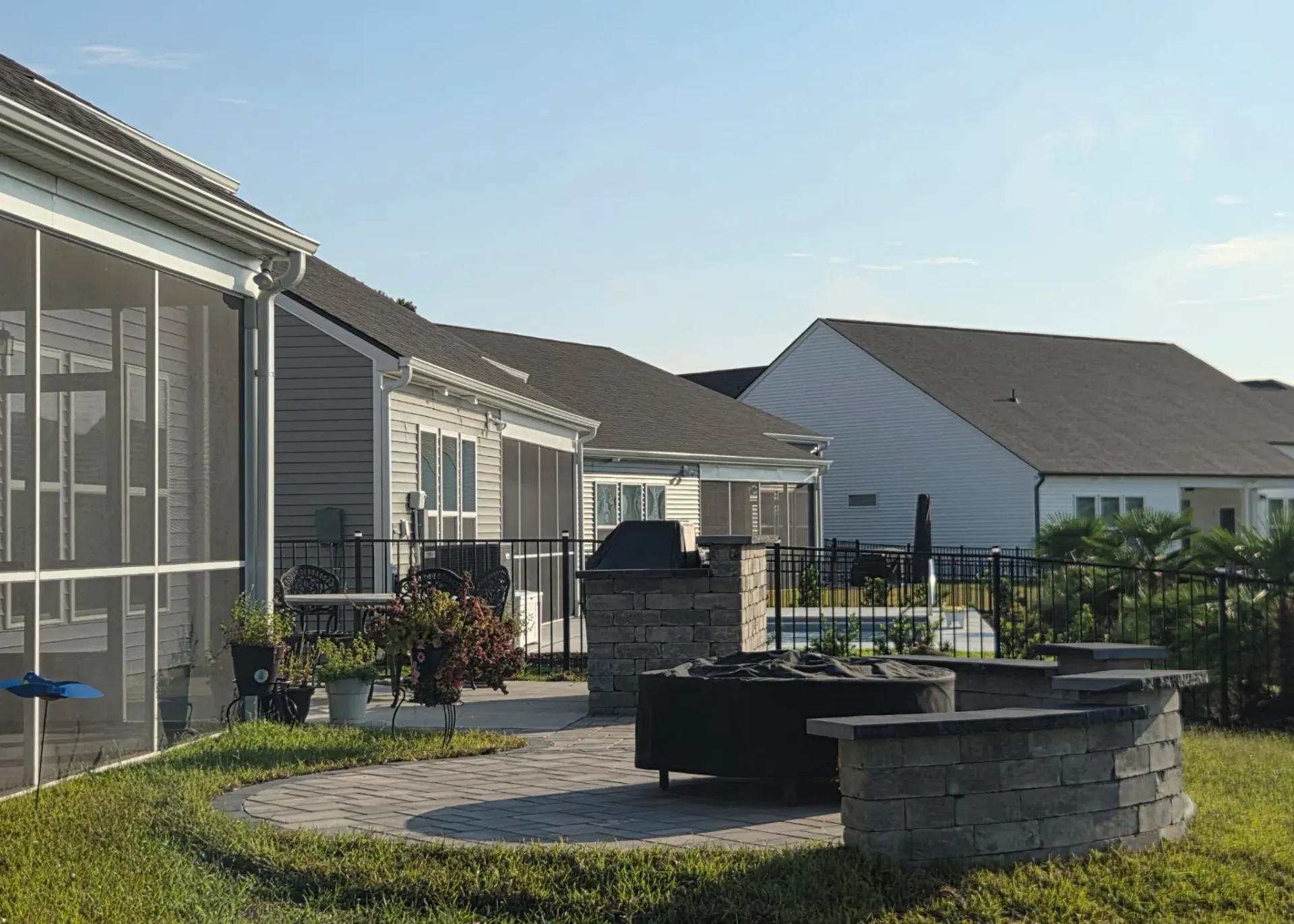 Suburban backyard with patio, hedge, and stone fire pit beside single-story homes under a blue sky