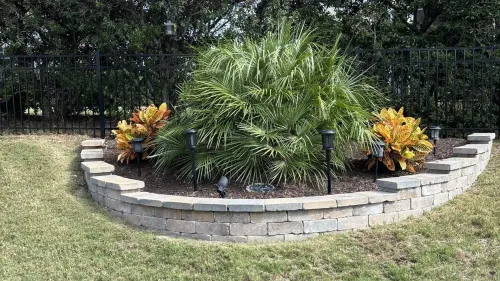 Circular garden bed with tropical plants and yellow-orange flowers bordered by stacked stone in a grassy yard