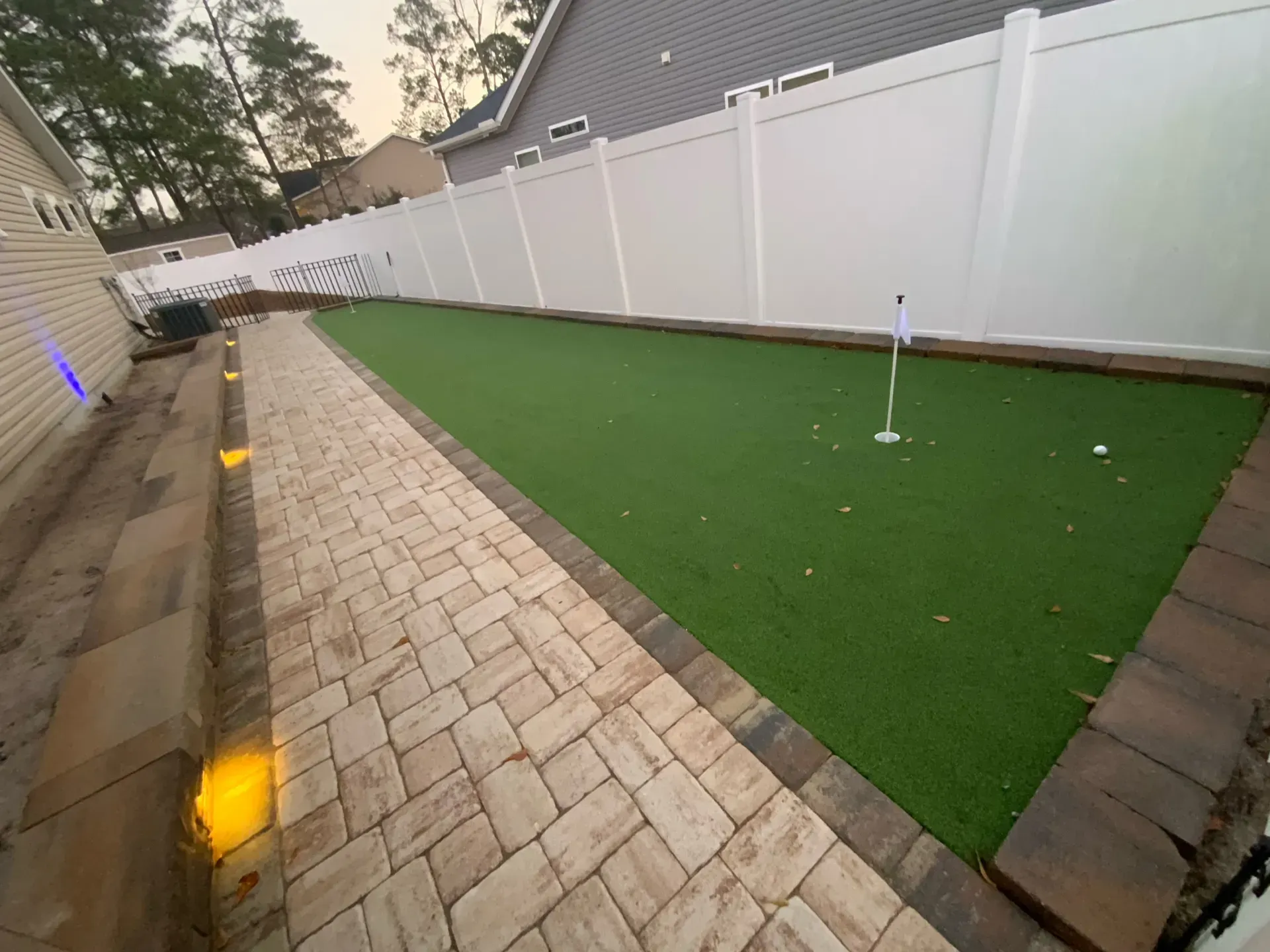 Backyard with white fence, paved walkway, and green artificial turf beside a house.