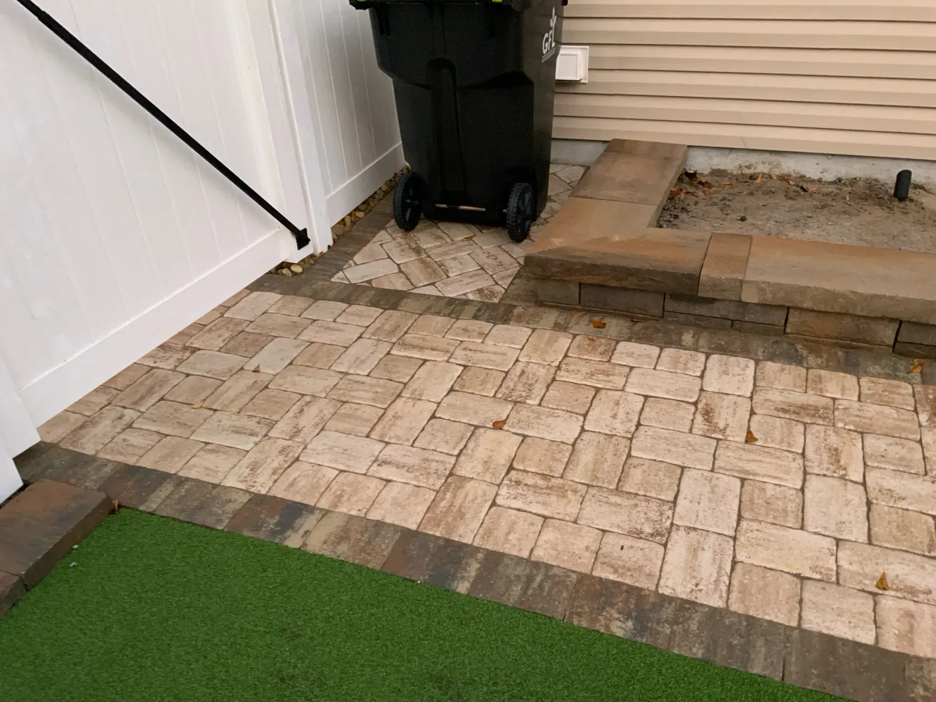 Stone patio with a black trash bin, wooden steps, and a strip of green artificial grass.