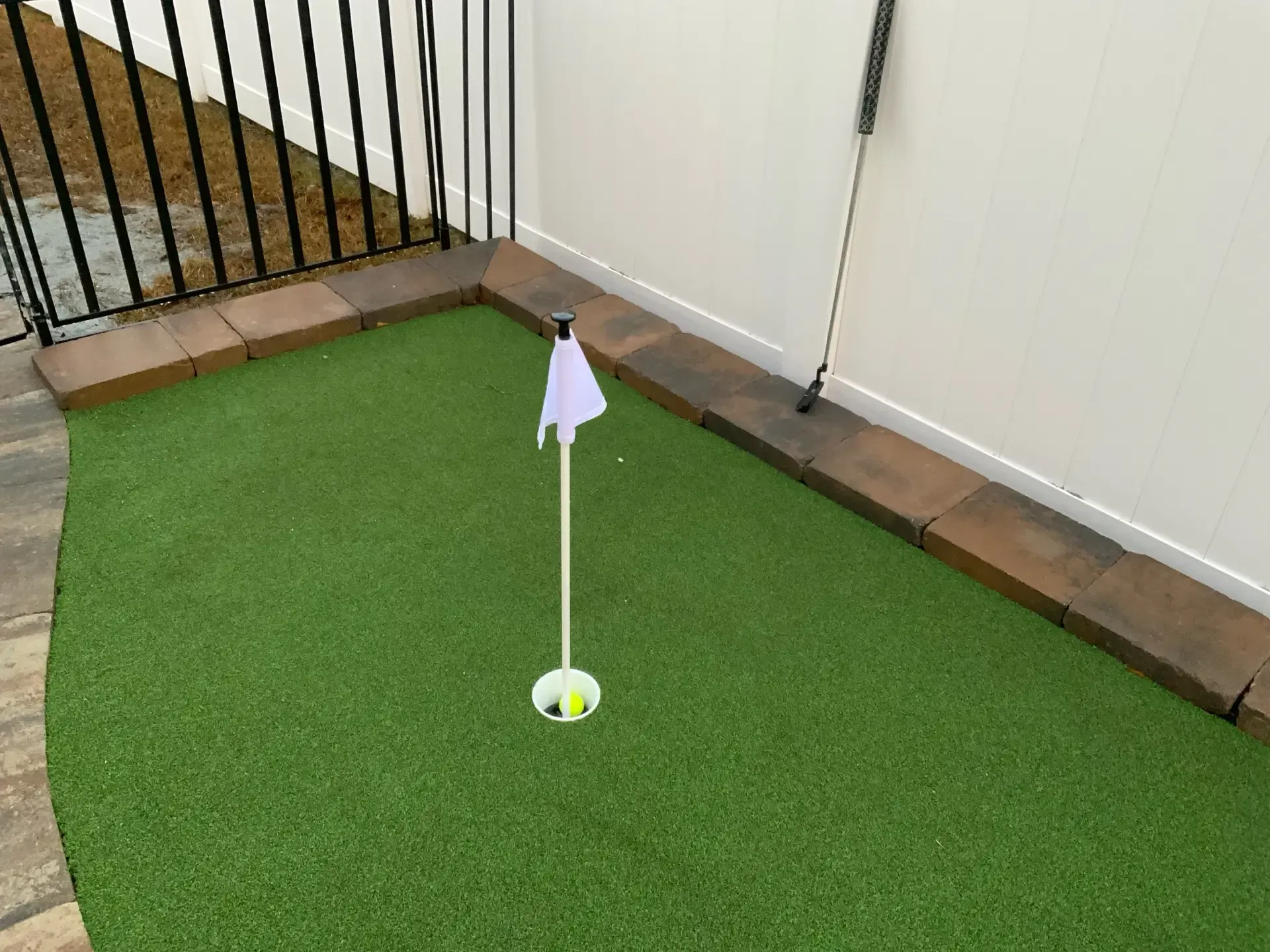 Golf hole with a white flag on green artificial turf beside a white fence and black railing